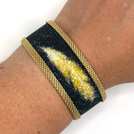 Close-up of wrist wearing a narrow cuff bracelet with matte black Swarovski micro-crystals surrounding a shimmery golden crystal feather image, with gold mesh edges, on a white background.