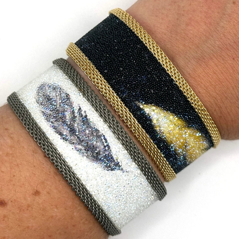 Close-up of wrist wearing two narrow cuff bracelets, one with matte black Swarovski micro-crystals surrounding a shimmery golden crystal feather image with gold mesh edges, the other with white Swarovski micro-crystals surrounding a shimmery black crystal feather image, with matte silver mesh edges, on a white background.