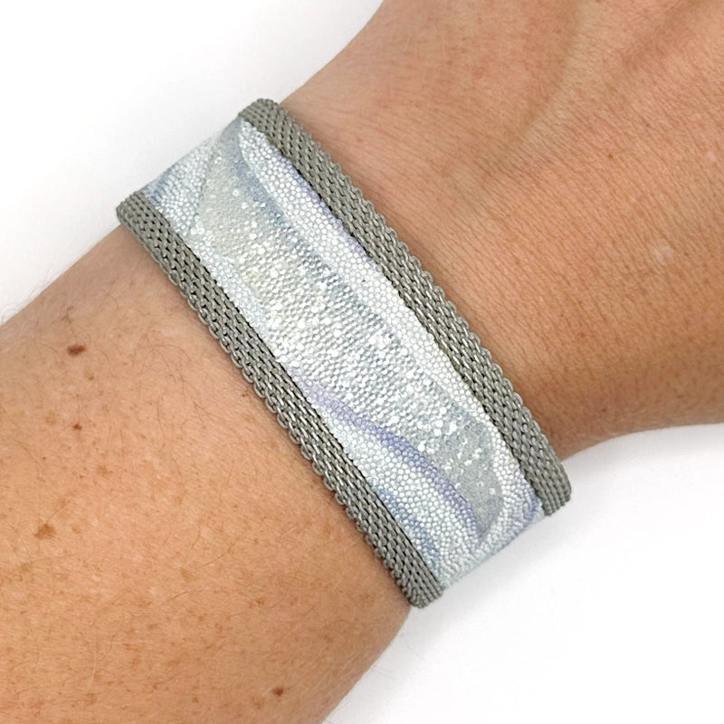 Close-up of wrist wearing a narrow cuff bracelet with blue lace agate slice pattern highlighted by shiny and matte Swarovski micro-crystals, with matte silver mesh edges, on a white background.