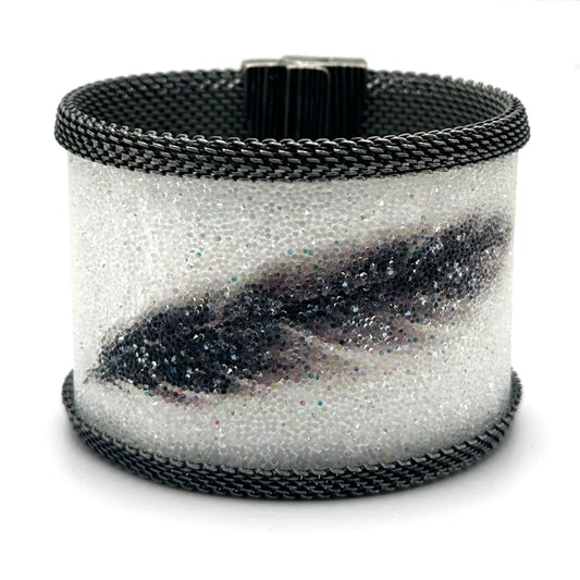 Wide cuff bracelet with white Swarovski micro-crystals surrounding a shimmery black crystal feather image, with gunmetal mesh edges, tilted to show the closed magnetic clasp, on a white background.