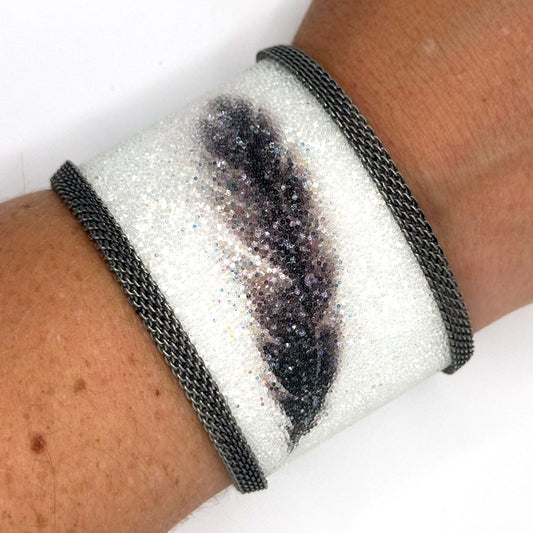 Closeup of wrist wearing a wide cuff bracelet with white Swarovski micro-crystals surrounding a shimmery black crystal feather image, with gunmetal mesh edges, on a white background.