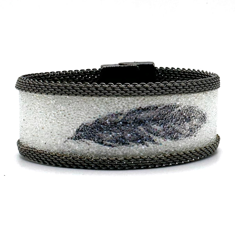 Narrow cuff bracelet with white Swarovski micro-crystals surrounding a shimmery black crystal feather image, with matte silver mesh edges, tilted to show the closed magnetic clasp, on a white background.