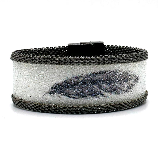Narrow cuff bracelet with white Swarovski micro-crystals surrounding a shimmery black crystal feather image, with matte silver mesh edges, tilted to show the closed magnetic clasp, on a white background.