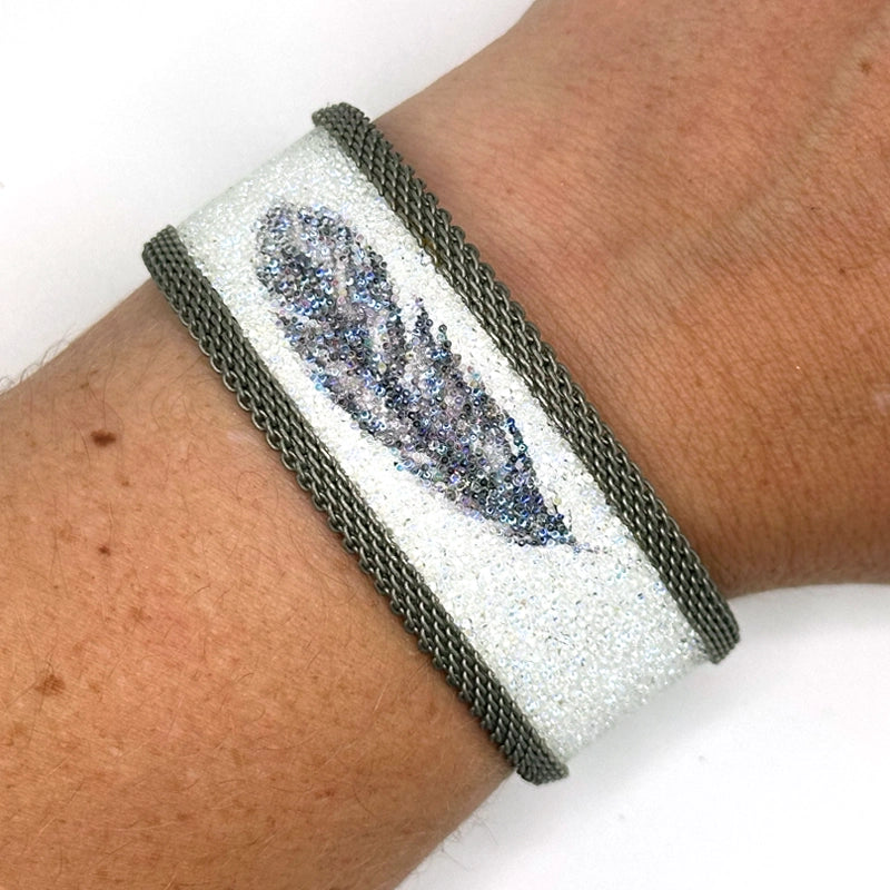 Close-up of wrist wearing a narrow cuff bracelet with white Swarovski micro-crystals surrounding a shimmery black crystal feather image, with matte silver mesh edges, on a white background.