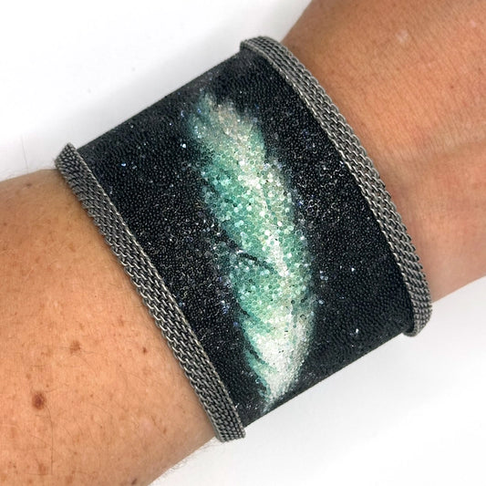 Closeup of wrist wearing a wide cuff bracelet with matte black Swarovski micro-crystals surrounding a shimmery green and white crystal feather image, with oxidized silver mesh edges, on a white background.
