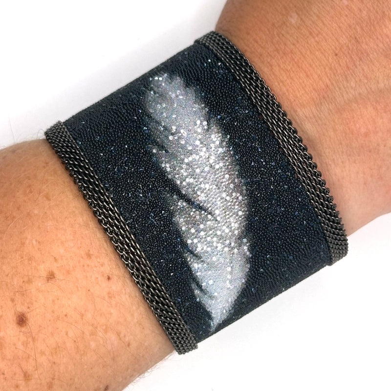 Closeup of wrist wearing a wide cuff bracelet with matte black Swarovski micro-crystals surrounding a shimmery white crystal feather image, with gunmetal mesh edges, on a white background.