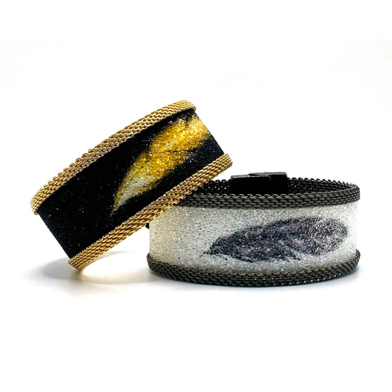 Two narrow cuff bracelets, one with matte black Swarovski micro-crystals surrounding a shimmery golden crystal feather image with gold mesh edges, the other with white Swarovski micro-crystals surrounding a shimmery black crystal feather image, with matte silver mesh edges,  on a white background.