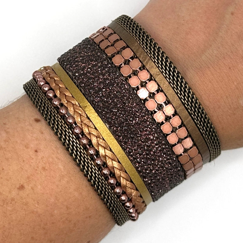Close-up of a wrist wearing a wide cuff bracelet with a mix of copper colored metal elements, metallic gold leather braids or strips, and pink-brown shimmery stingray, with brass mesh edges, on a white background.