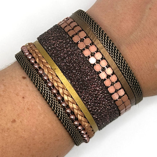 Close-up of a wrist wearing a wide cuff bracelet with a mix of copper colored metal elements, metallic gold leather braids or strips, and pink-brown shimmery stingray, with brass mesh edges, on a white background.