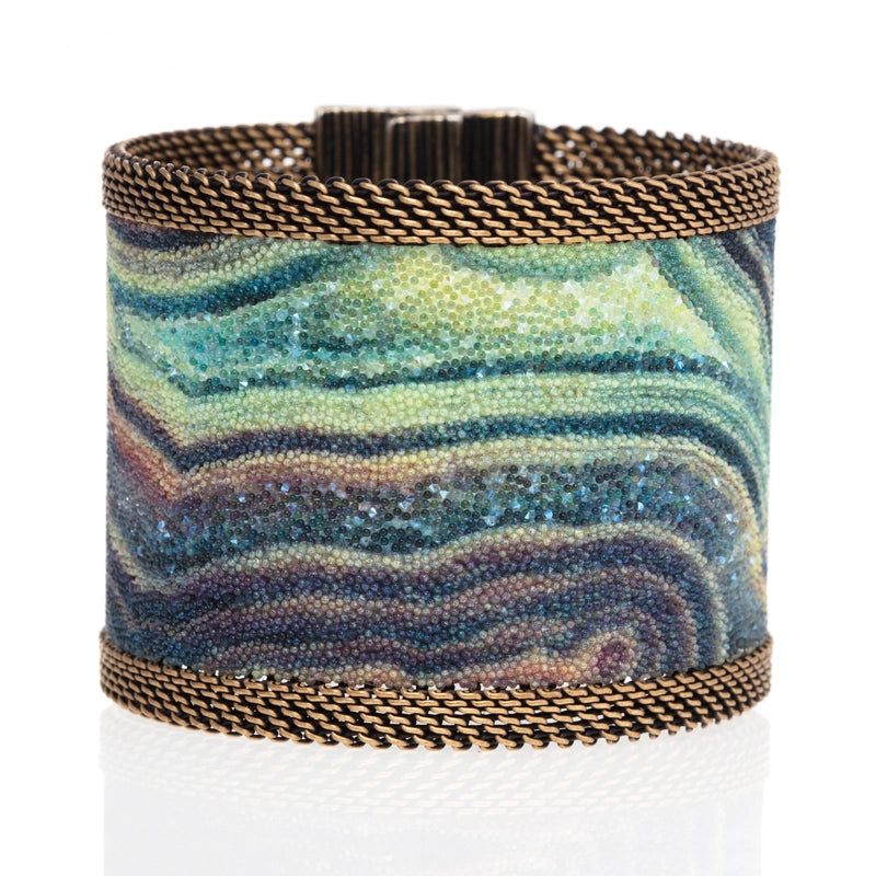 Wide cuff bracelet with multi-color agate slice pattern highlighted by shiny and matte Swarovski micro-crystals, with brass mesh edges, tilted to show closed magnetic clasp, on a white background.