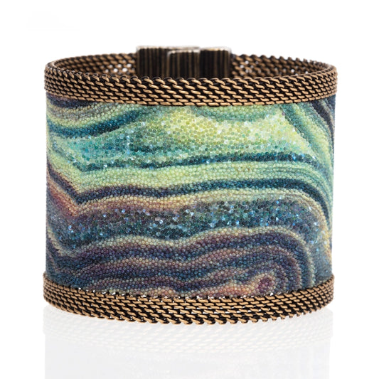 Wide cuff bracelet with multi-color agate slice pattern highlighted by shiny and matte Swarovski micro-crystals, with brass mesh edges, tilted to show closed magnetic clasp, on a white background.