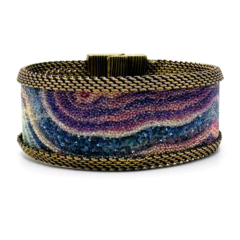 Narrow cuff bracelet with purple-blue multi-color agate slice pattern highlighted by shiny and matte Swarovski micro-crystals, with brass mesh edges, on a white background.