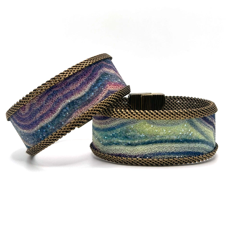 Two narrow cuff bracelets with multi-color agate slice patterns highlighted by shiny and matte Swarovski micro-crystals, with brass mesh edges, on a white background.