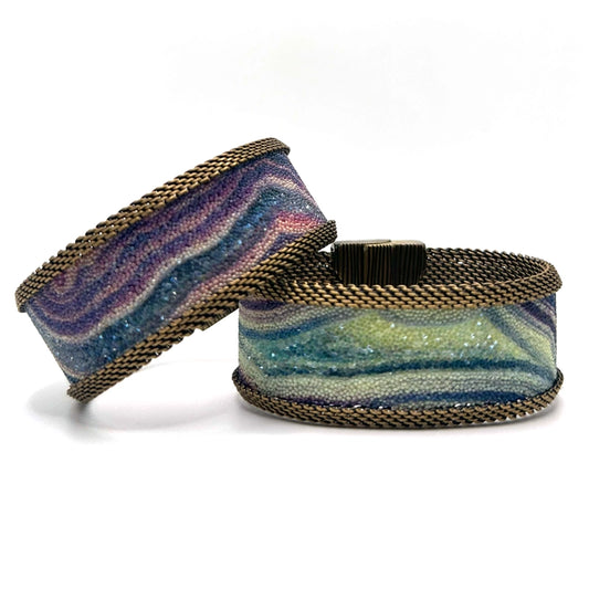 Two narrow cuff bracelets with multi-color agate slice patterns highlighted by shiny and matte Swarovski micro-crystals, with brass mesh edges, on a white background.