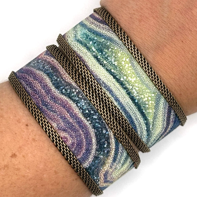 Closeup of a wrist wearing two narrow cuff bracelets with multi-color agate slice patterns highlighted by shiny and matte Swarovski micro-crystals, with brass mesh edges, on a white background.